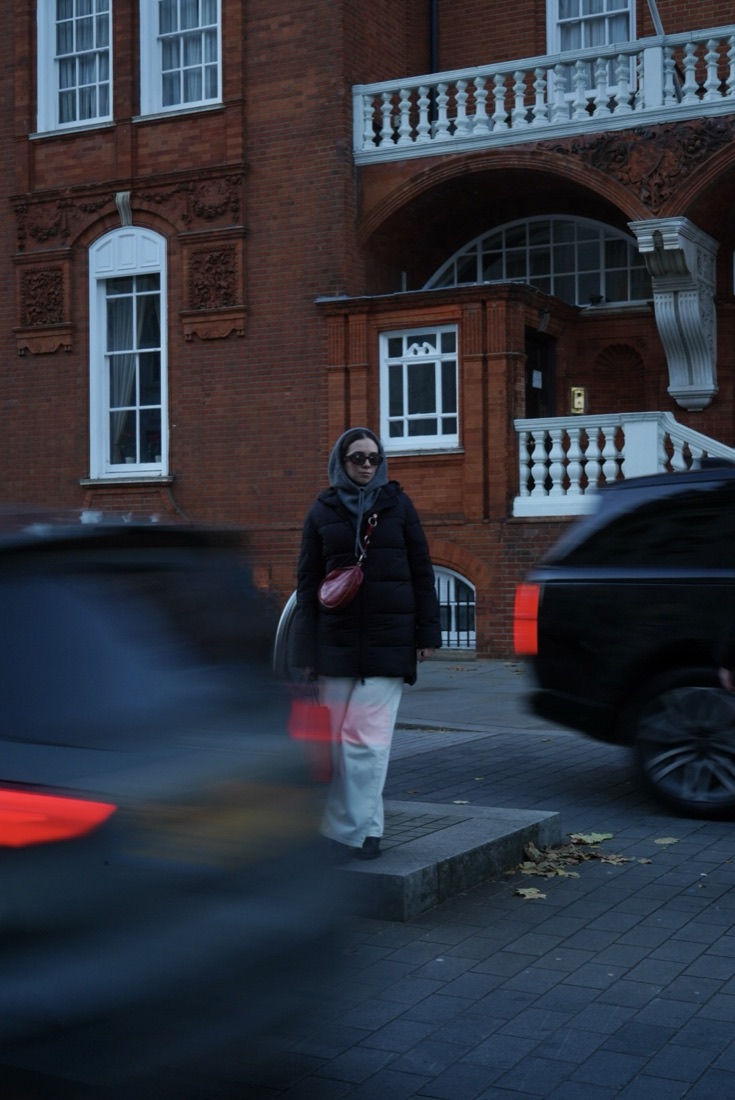Woman in a puffer jacket by a red-brick building with traffic blurring past