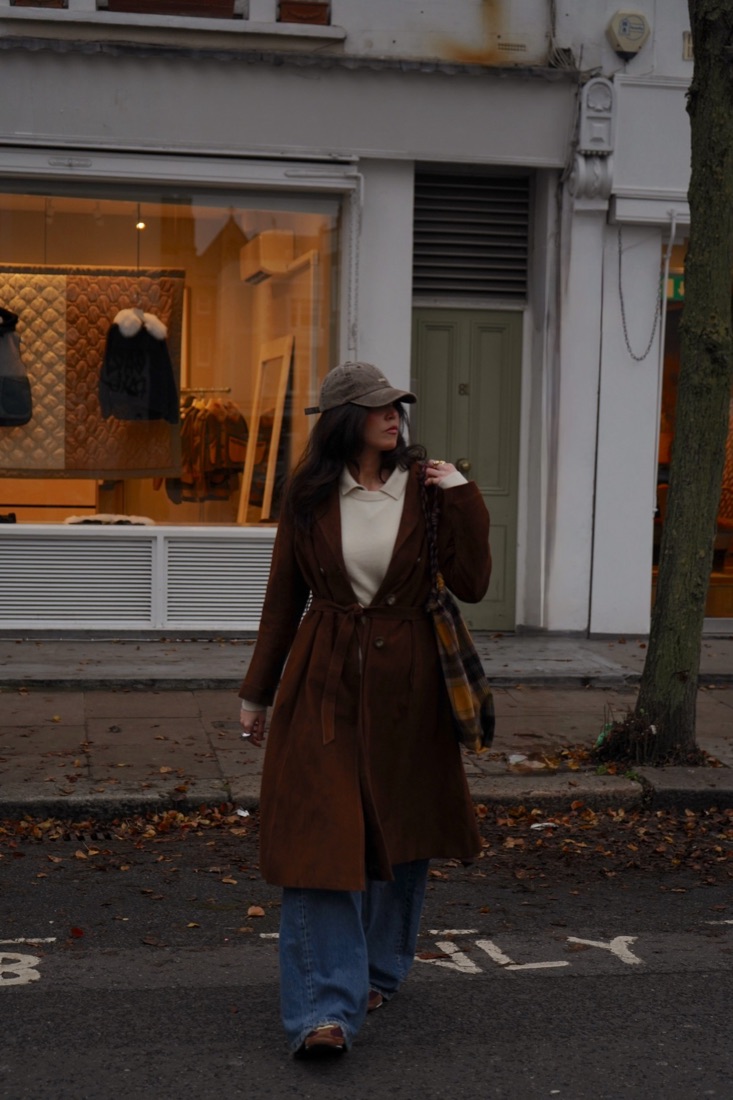 Woman in a brown coat crossing the street in front of a lit shop window